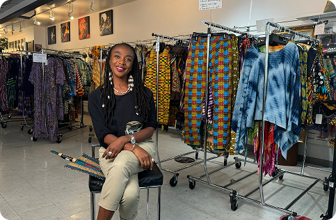 A professional photo of Keithy Antoine, a Black woman with long, braided hair adorned with shells. She is sitting on a chair, smiling at the camera in the middle of a clothing store. She is holding a colorful, woven fan. The store is filled with racks of clothing featuring vibrant patterns and textures.